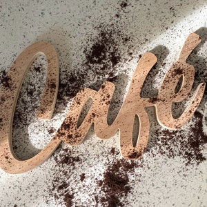 May include: A wooden sign that says "Cafe" in a cursive font. The sign is covered in brown coffee grounds.