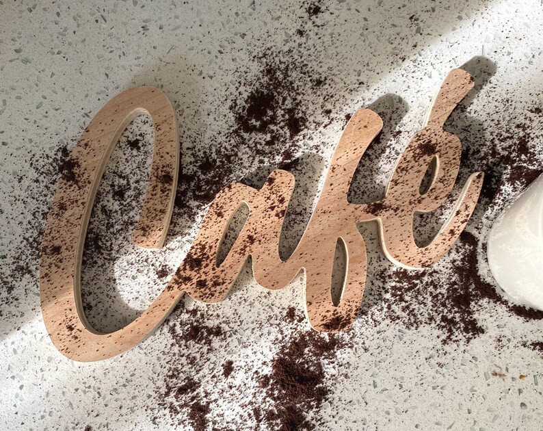 May include: A wooden sign that says "Cafe" in a cursive font. The sign is covered in brown coffee grounds.