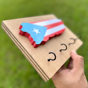 K&ouml;nnte beinhalten: Schl&uuml;sselhalter aus Holz in Form der Umrisse von Puerto Rico, bemalt in den Farben der puerto-ricanischen Flagge: Rot, Wei&szlig; und Blau. Der Schl&uuml;sselhalter hat drei Metallhaken.