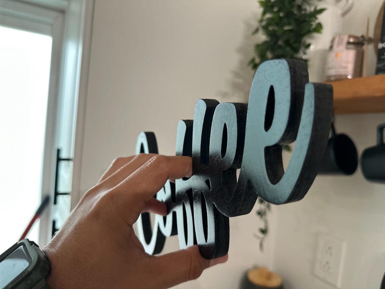 May include: Black wooden sign spelling "Coffee" in cursive. The sign is held by a hand. A decorative item for a kitchen or coffee bar. The sign is made of wood.