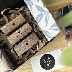 Puede incluir: Una caja de regalo llena de virutas de papel marr&oacute;n y tres bloques de madera apilados uno encima del otro. Los bloques tienen la palabra "Recuerdos" grabada en ellos con un s&iacute;mbolo de coraz&oacute;n. Una peque&ntilde;a tarjeta con el texto "Gracias" y un s&iacute;mbolo de coraz&oacute;n est&aacute; sobre la mesa junto a la caja.