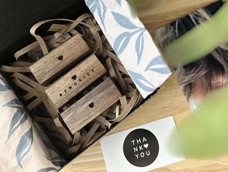 Puede incluir: Una caja de regalo llena de virutas de papel marr&oacute;n y tres bloques de madera apilados uno encima del otro. Los bloques tienen la palabra "Recuerdos" grabada en ellos con un s&iacute;mbolo de coraz&oacute;n. Una peque&ntilde;a tarjeta con el texto "Gracias" y un s&iacute;mbolo de coraz&oacute;n est&aacute; sobre la mesa junto a la caja.