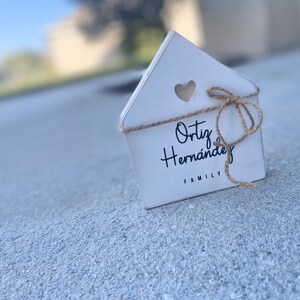 May include: White wooden house-shaped sign with a heart cutout and a twine bow. The sign reads "Ortiz Hernandez Family".