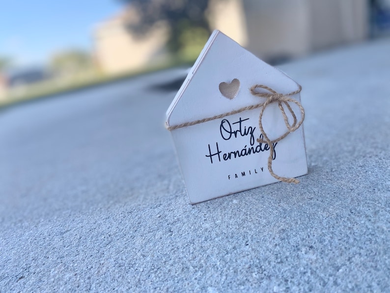 May include: White wooden house-shaped sign with a heart cutout and a twine bow. The sign reads "Ortiz Hernandez Family".