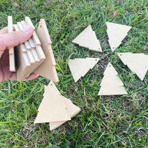 May include: A set of nine unfinished wooden Christmas tree decorations. The decorations are in the shape of a triangle and are arranged in a circle on a bed of green grass.