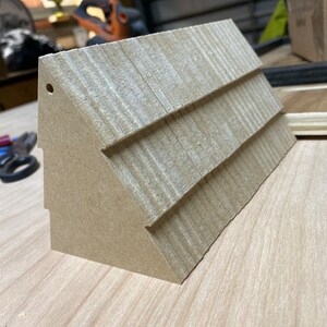 May include: A small, brown, wooden model of a house with a shingled roof. The model has a hole in the top and is sitting on a wooden surface.