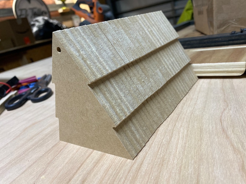 May include: A small, brown, wooden model of a house with a shingled roof. The model has a hole in the top and is sitting on a wooden surface.
