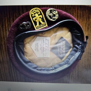 May include: A maroon wool beret featuring a black leather trim and three metallic pins. The pins include a yellow and black patch with the text 509 GERONIMO, a circular emblem, and a dark winged insignia. The interior lining is tan with a label.