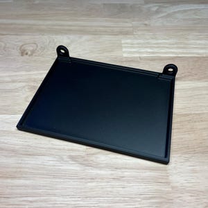 May include: A square, black metal tray with a raised edge and two small, circular holes on opposite corners. The tray is designed to hold small items and has a matte finish. The tray is shown on a light wood surface.
