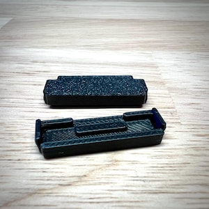 May include: Two small, dark teal-colored rectangular objects with textured surfaces. One object is a solid rectangle, while the other has a recessed area and raised sections. The objects are on a light wood-grain surface.