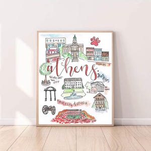 May include: Watercolour print depicting Athens, Georgia, with key landmarks and the city's name in cursive script. The artwork includes illustrations of buildings, a tree, and a football stadium, framed in wood.
