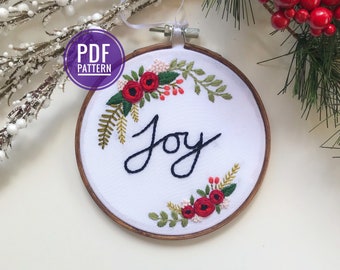 Peut inclure: Un cercle de broderie blanc avec un cadre en bois, orné d'un motif floral rouge et du mot "Joie" brodé en fil noir.