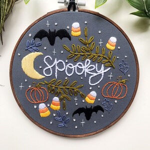 May include: Embroidered hoop art with a gray background featuring the word "spooky" in white cursive lettering. The design includes bats, pumpkins, candy corn, and leaves in various colors.