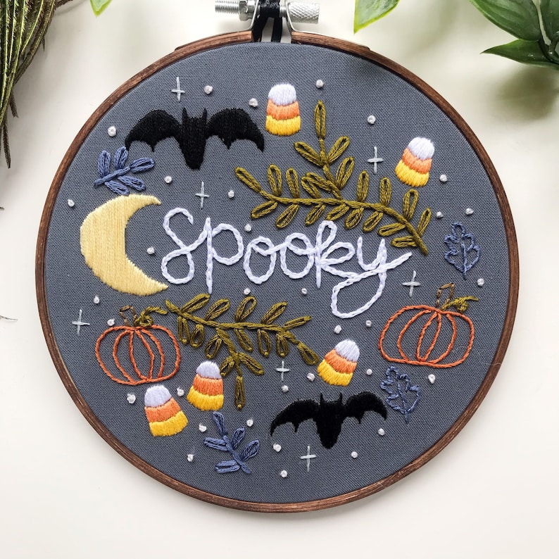 May include: Embroidered hoop art with a gray background featuring the word "spooky" in white cursive lettering. The design includes bats, pumpkins, candy corn, and leaves in various colors.
