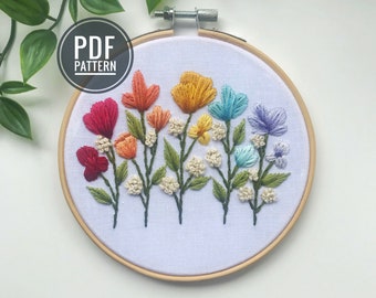 May include: Embroidered hoop art featuring a rainbow of flowers. The flowers are stitched in red, orange, yellow, green, blue, and purple. The hoop is made of wood and has a white fabric background.