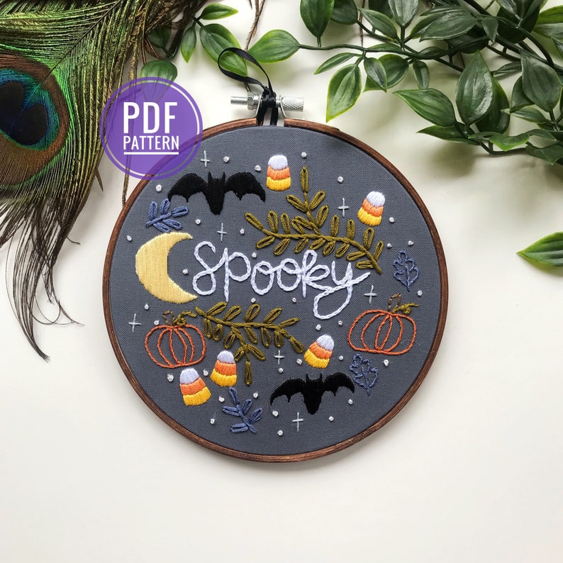 May include: Embroidered hoop art with the word "spooky" in white script surrounded by bats, pumpkins, candy corn, and leaves on a gray background.