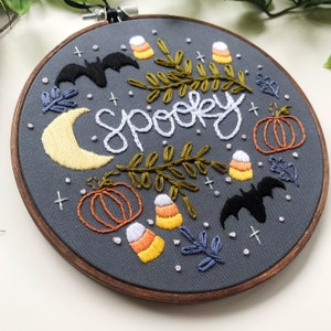 May include: Embroidered hoop art with the word "spooky" in white thread on a gray background. The design includes bats, pumpkins, candy corn, and leaves in various colors.