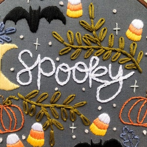 May include: Embroidered hoop art with the word "Spooky" in white thread, surrounded by bats, candy corn, pumpkins, and leaves in shades of yellow, orange, and green on a gray background.