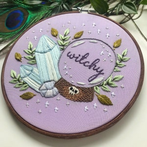May include: Embroidered hoop art with a purple background featuring a crystal ball with the word "witchy" inside. The crystal ball is surrounded by green leaves, blue crystals, and gold leaves. There are white stars scattered around the hoop.