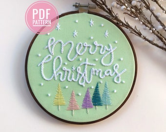 Könnte beinhalten: Ein grüner Stickrahmen mit weißer Stickerei, auf dem "Frohe Weihnachten" steht, und fünf bunte gestickte Weihnachtsbäume.