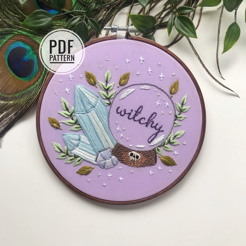 May include: A purple embroidery hoop with a crystal, leaves, and a glass orb with the word "witchy" inside. The hoop is on a white surface.