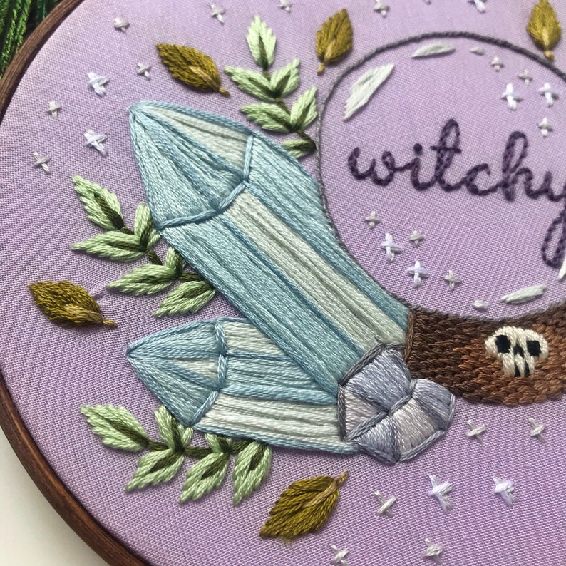 May include: Embroidered hoop art with a purple background featuring two blue crystals, green leaves, and a brown crystal ball with the word "witchy" embroidered on it.