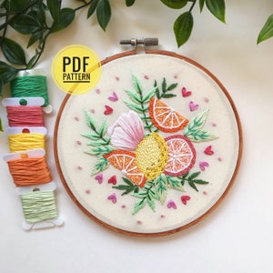 以下が含まれることがあります： レモン、オレンジ、ピンクの花をモチーフにした刺繍フープのPDFパターン。デザインは緑の葉とピンクのハートで囲まれています。