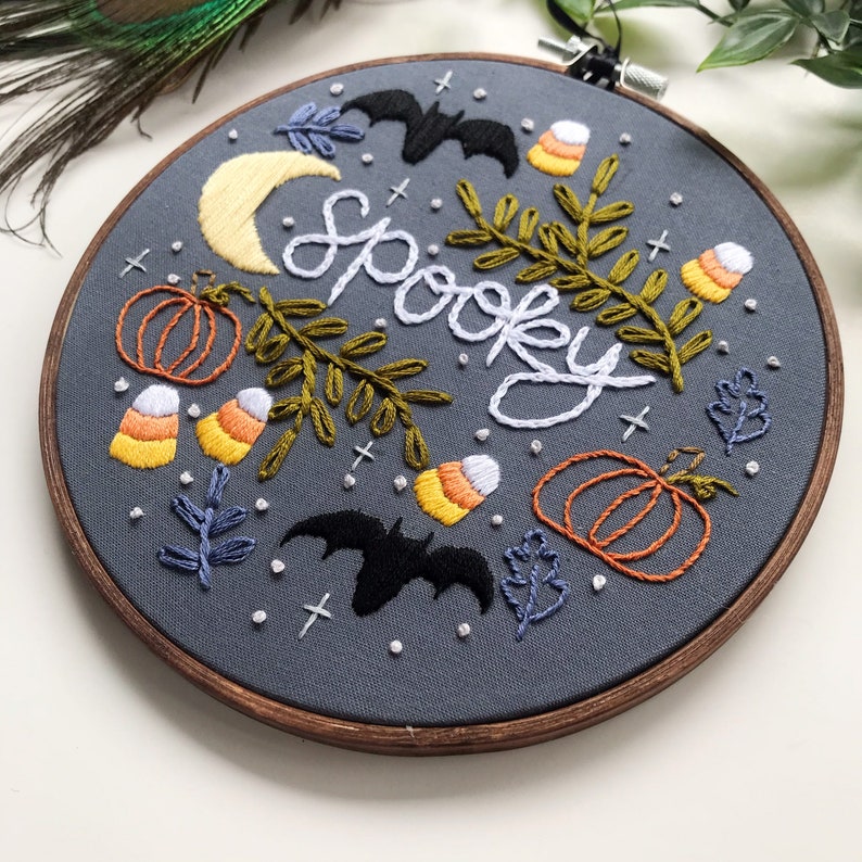 May include: Embroidered hoop art with a gray background featuring the word "Spooky" in white thread, surrounded by bats, pumpkins, candy corn, and leaves in various colors.
