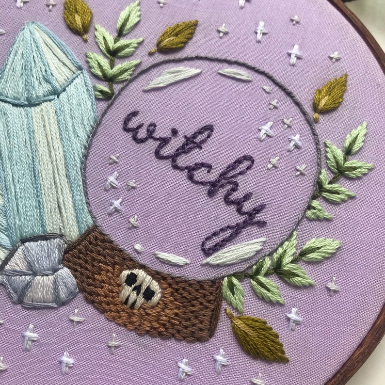 May include: Embroidered hoop art with a purple background, a crystal, leaves, and a circle with the word "Witchy" inside.