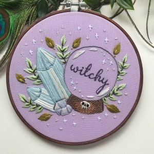 May include: Embroidered hoop art with a purple background featuring a crystal, leaves, and a glass orb with the word "witchy" inside.