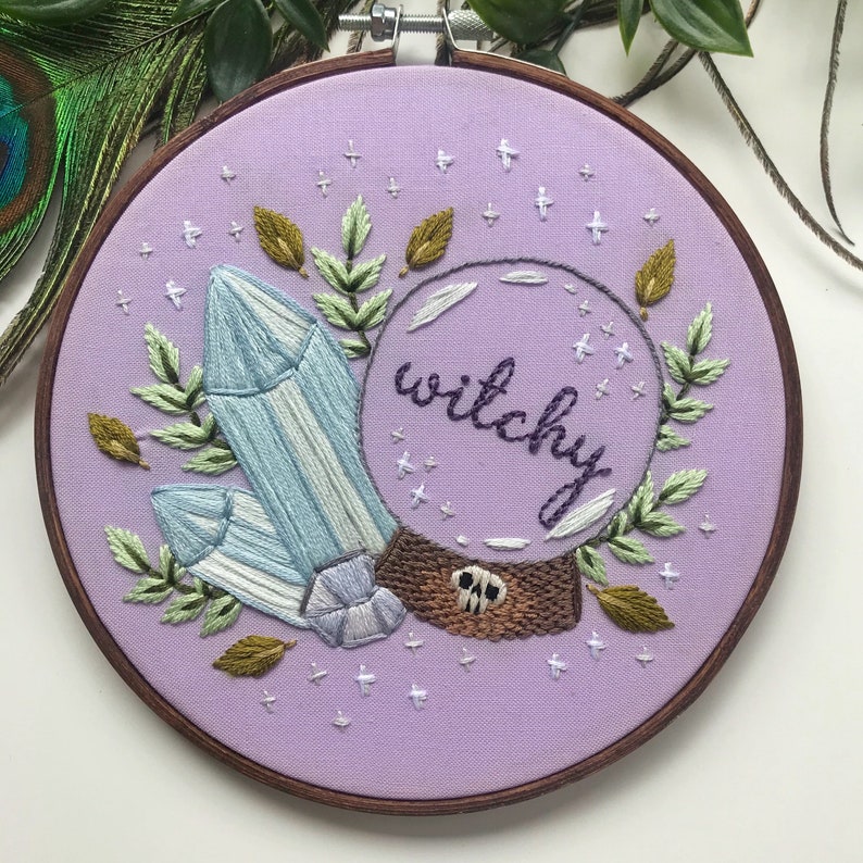 May include: Embroidered hoop art with a purple background featuring a crystal, leaves, and a glass orb with the word "witchy" inside.