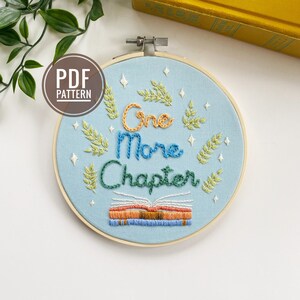May include: A blue embroidery hoop featuring the text "One More Chapter" in colourful cursive lettering. The text is surrounded by green leaves and white stars. A stack of books is embroidered below the text.
