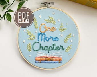 以下が含まれることがあります： 青い刺繍枠に、「One More Chapter」というカラフルな筆記体で書かれた文字が刺繍されています。文字の周りは緑の葉と白い星で囲まれています。文字の下には、本の束が刺繍されています。