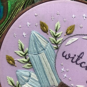 May include: Embroidered hoop art with a purple background featuring two light blue crystals, green leaves, and gold leaves. The text "wild" is embroidered in purple.
