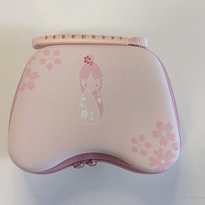 Puede incluir: Estuche de transporte rosa con cierre de cremallera y asa. El estuche tiene un dise&ntilde;o floral con una peque&ntilde;a mu&ntilde;eca en el centro.