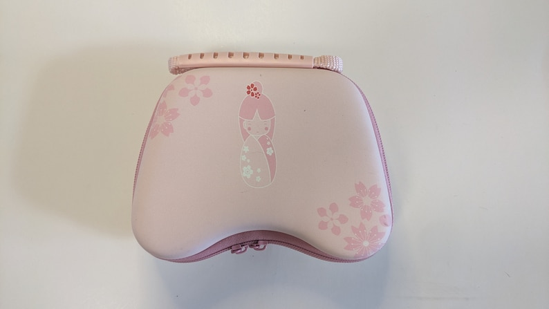 Puede incluir: Estuche de transporte rosa con cierre de cremallera y asa. El estuche tiene un dise&ntilde;o floral con una peque&ntilde;a mu&ntilde;eca en el centro.