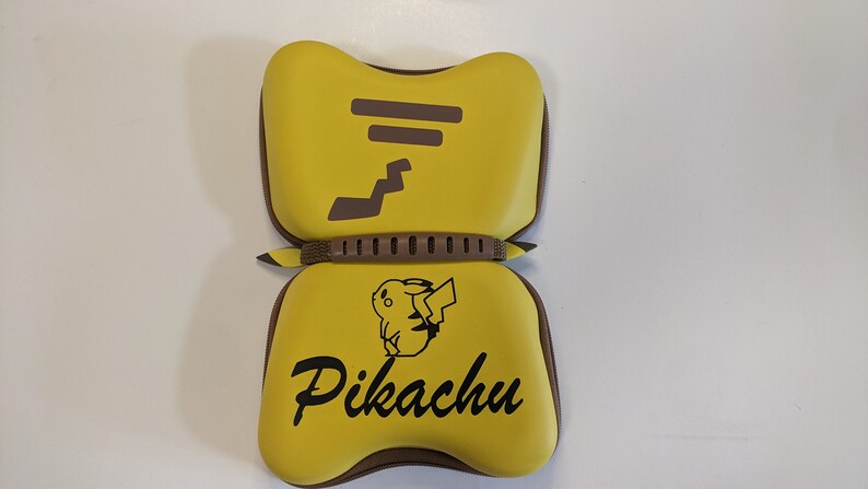 Puede incluir: Estuche de transporte amarillo y marr&oacute;n para Nintendo Switch con un dise&ntilde;o de Pikachu. El estuche tiene una cremallera y un asa.
