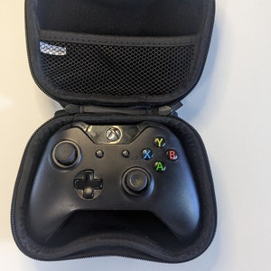 Puede incluir: Un mando Xbox negro en una funda de transporte negra con cremallera. El mando tiene un logotipo de Xbox negro y verde en la parte superior y botones negros con letras de colores: A, B, X e Y.