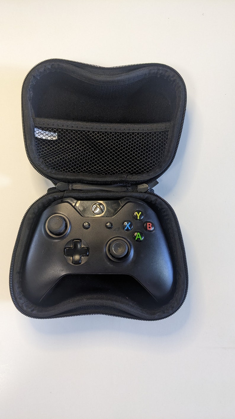 Puede incluir: Un mando Xbox negro en una funda de transporte negra con cremallera. El mando tiene un logotipo de Xbox negro y verde en la parte superior y botones negros con letras de colores: A, B, X e Y.