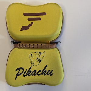 May include: A yellow carrying case with a brown handle and a Pikachu design. The case has a zipper closure and a brown trim.
