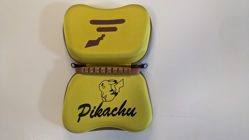 Puede incluir: Una funda de transporte amarilla con un asa marr&oacute;n y un dise&ntilde;o de Pikachu. La funda tiene una cremallera y un ribete marr&oacute;n.