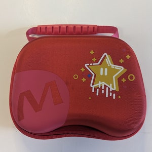 Puede incluir: Estuche de transporte rojo con una estrella y una M en la parte delantera. El estuche tiene un asa en la parte superior.