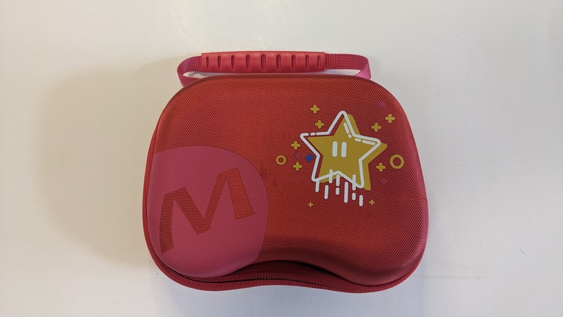 Puede incluir: Estuche de transporte rojo con una estrella y una M en la parte delantera. El estuche tiene un asa en la parte superior.