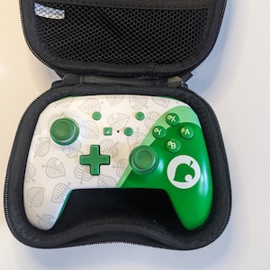 Puede incluir: Un mando de Nintendo Switch blanco y verde con un patr&oacute;n de hojas y el logotipo de Animal Crossing. El mando est&aacute; en una funda de transporte negra.