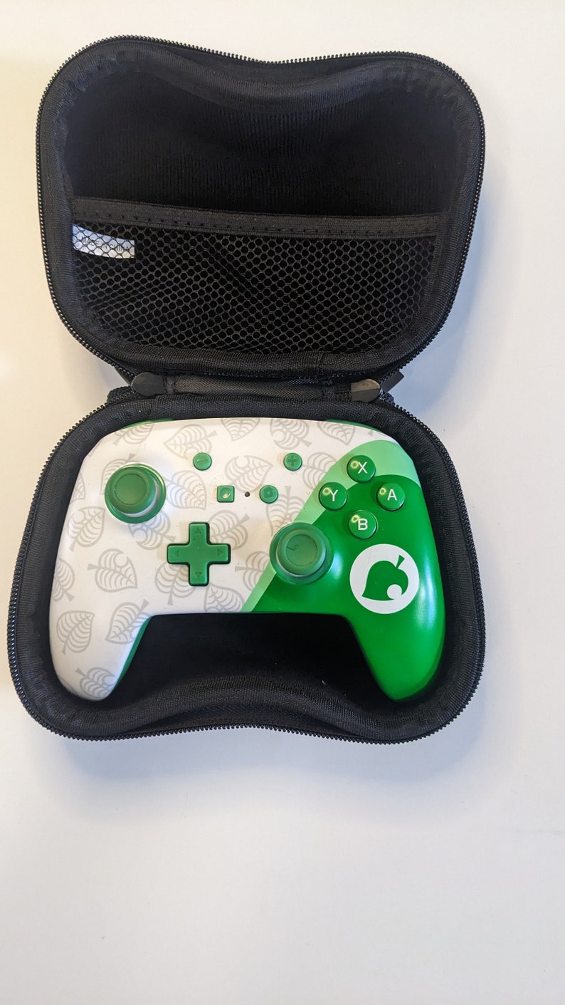 Puede incluir: Un mando de Nintendo Switch blanco y verde con un patr&oacute;n de hojas y el logotipo de Animal Crossing. El mando est&aacute; en una funda de transporte negra.