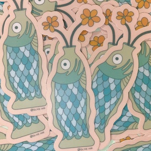 May include: A stack of stickers featuring a green fish with blue scales holding a flower in its mouth. The fish is in a vase with a green stem and yellow flowers. The stickers have the text "@OLIVE_INK" on them.