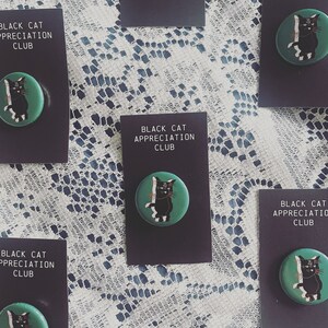 May include: A set of six black cat appreciation club pins. Each pin features a black cat with white markings on a teal green background. The pins are displayed on black card stock with the text "Black Cat Appreciation Club".