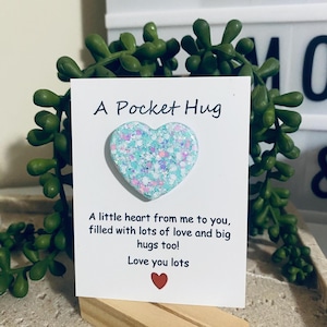 May include: A Pocket Hug card featuring a heart-shaped decoration. The heart is light blue with glitter. The card reads "A Pocket Hug" and includes a message about love and hugs. A small red heart is at the bottom of the card.