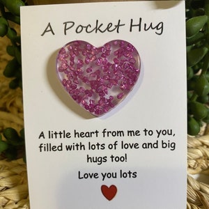 May include: A white card with the text "A Pocket Hug" and a pink heart-shaped resin charm filled with glitter. The card also includes the text "A little heart from me to you, filled with lots of love and big hugs too! Love you lots" and a small red heart.