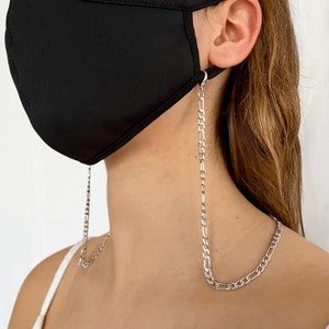 May include: A silver chain mask holder is shown, attached to a black face mask. The chain has a flat, linked design. The mask is made of a smooth, dark fabric. The background is a plain, light color.